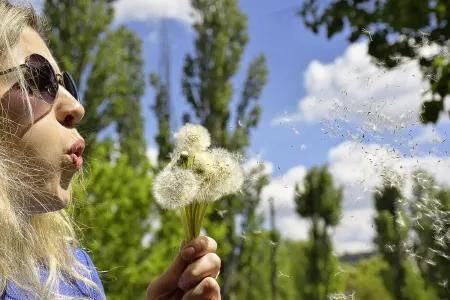 Junge Frau bläst Löwenzahnsamen in die Luft an einem sonnigen Tag – symbolisch für Frühling, Natur und Pollenflug.