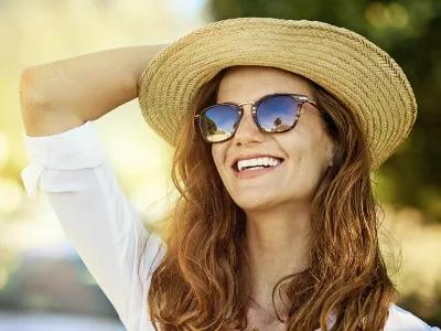 Lächelnde Frau mit Strohhut und Sonnenbrille im Freien. Sie trägt ein weißes Hemd und hält sich mit einer Hand am Hut fest. Die Szene vermittelt Sommer, Sonnenschutz und gute Laune.