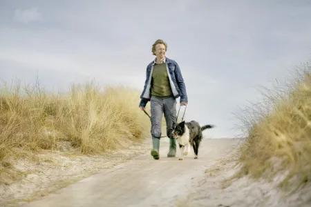 Älterer Mann spaziert mit seinem Hund an einem Dünenweg entlang – Bewegung an der frischen Luft als wichtiger Bestandteil eines gesunden Lebensstils bei Diabetes.