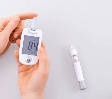 Hand hält ein digitales Blutzuckermessgerät mit sichtbarem Wert, daneben liegt ein Stechhilfegerät – Darstellung von klassischer Blutzuckermessung bei Diabetes.