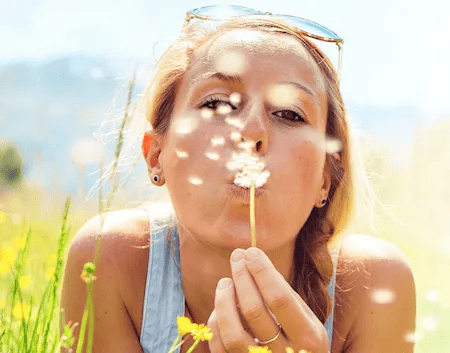 Junge Frau mit Sonnenbrille im Haar pustet eine Pusteblume in die Kamera. Sie befindet sich auf einer blühenden Sommerwiese mit Bergen im Hintergrund. Die Szene vermittelt Leichtigkeit, Natur und Frühlingsgefühle.