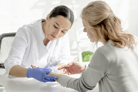 Eine Hautexpertin mit blauen Einmalhandschuhen untersucht die Hand einer Frau in heller Umgebung. Die Szene vermittelt professionelle Beratung und Hautpflege im medizinischen Kontext.
