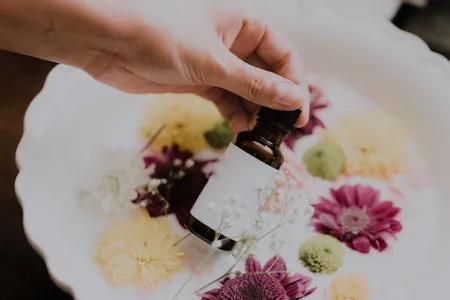 Hand hält eine braune Tropfflasche über einer Schale mit Wasser und bunten Blüten – Darstellung von natürlichem Insektenschutz oder ätherischen Ölen.