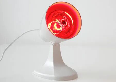 Wei&szlig;e Infrarotlampe mit rotem Licht leuchtet zur W&auml;rmebehandlung, auf einem Tisch vor wei&szlig;em Hintergrund.