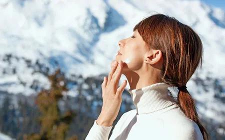 Frau im Profil mit hochgestecktem Haar trägt einen weißen Rollkragenpullover und cremt ihr Gesicht bei Sonnenschein in einer winterlichen Berglandschaft ein.