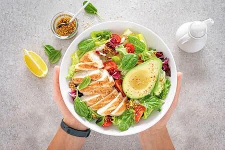 Frischer bunter Salat mit gegrillter Hähnchenbrust und einer halben Avocado, angerichtet in einer weißen Schale und gehalten von zwei Händen, daneben Zitrone und Dressing.
