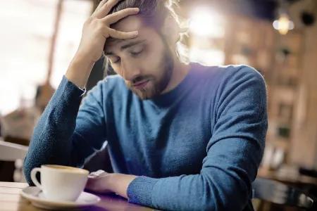 Ein junger Mann mit langen Haaren in einem blauen Pullover sitzt ersch&ouml;pft in einem Caf&eacute;, st&uuml;tzt den Kopf auf eine Hand und blickt niedergeschlagen auf eine Tasse Kaffee vor ihm.