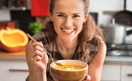 Eine lachende Frau h&auml;lt in der K&uuml;che eine Sch&uuml;ssel mit K&uuml;rbissuppe und einen L&ouml;ffel in der Hand &ndash; Symbol f&uuml;r gesunde, warme Ern&auml;hrung.