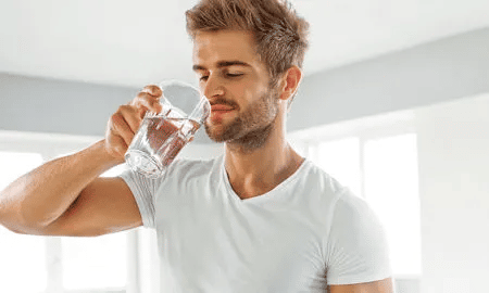 Ein junger Mann in wei&szlig;em T-Shirt trinkt ein Glas Wasser und schaut zufrieden &ndash; Symbol f&uuml;r gesunde Fl&uuml;ssigkeitszufuhr.