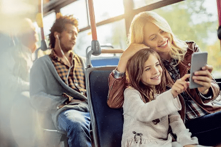 Eine Mutter und ihre Tochter sitzen in einem Bus und l&auml;cheln beim Blick auf ein Smartphone, ein anderer Fahrgast h&ouml;rt Musik im Hintergrund
