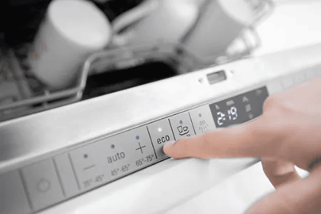 Eine Person dr&uuml;ckt die Eco-Taste an einem modernen Geschirrsp&uuml;ler, im Hintergrund sind gesp&uuml;lte Tassen sichtbar.