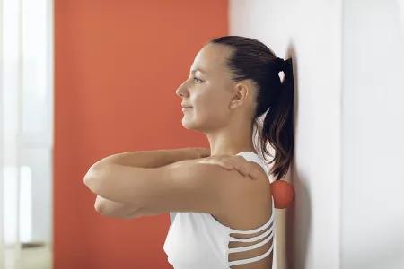 Eine Frau lehnt mit dem R&uuml;cken an einer Wand, wobei ein roter Faszienball zwischen ihrer Schulter und der Wand platziert ist. Sie tr&auml;gt ein wei&szlig;es Sporttop und hat die Arme vor der Brust verschr&auml;nkt.