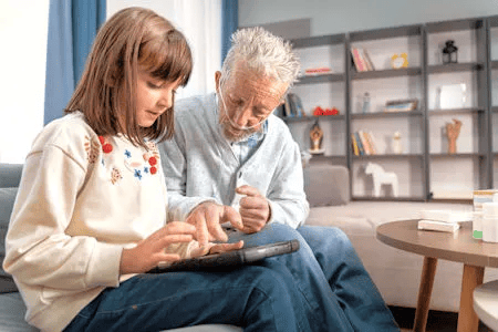 Ein &auml;lterer Mann mit grauen Haaren, Bart und Sauerstoffbrille sitzt auf einem Sofa neben einem jungen M&auml;dchen. Beide schauen konzentriert auf ein Tablet, das sie gemeinsam bedienen.