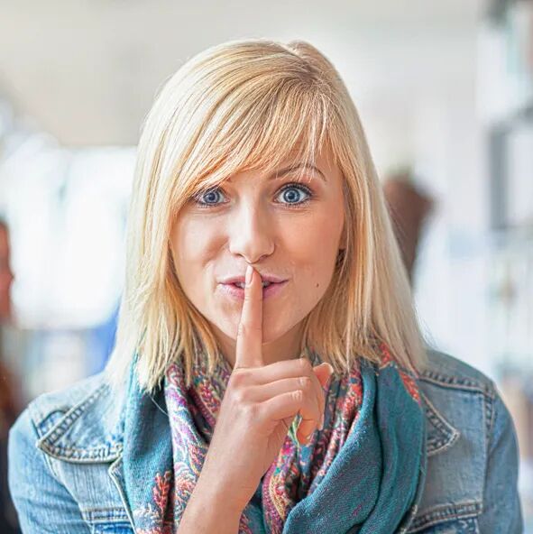 Eine junge blonde Frau mit Schal und Jeansjacke schaut direkt in die Kamera und h&auml;lt den Zeigefinger vor den Mund als Zeichen f&uuml;r Ruhe oder Geheimhaltung.