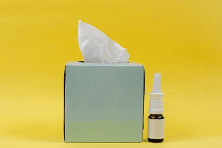 Taschentücherbox und Nasenspray vor gelbem Hintergrund als Symbol für Erkältung oder Grippe.