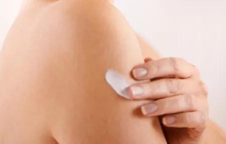 Eine Person cremt ihren Oberarm mit einer weißen Lotion ein. Die Haut ist hell, der Fokus liegt auf der Hand und dem eingecremten Bereich.