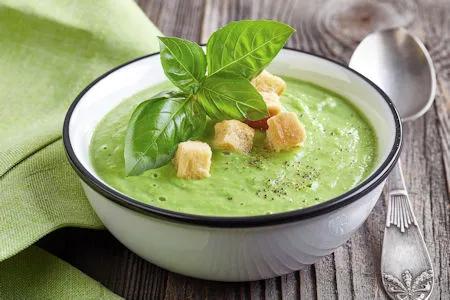 Cremige gr&uuml;ne Brokkoli-Erbsen-Cremesuppe mit Croutons und Basilikumbl&auml;ttern in einer wei&szlig;en Sch&uuml;ssel auf einem rustikalen Holztisch.