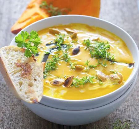 K&uuml;rbissuppe mit ger&ouml;steten K&uuml;rbiskernen, frischer Kresse und Brotscheibe in wei&szlig;er Schale serviert.