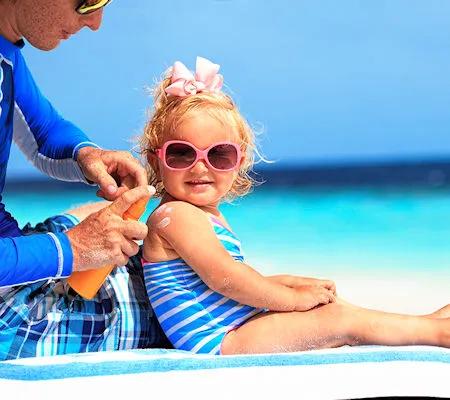 Ein kleines blondes Mädchen mit rosa Sonnenbrille und rosa Schleife sitzt im Badeanzug auf einem Badetuch am Strand, während ein Erwachsener ihr Sonnenschutzmittel aufträgt. Der Hintergrund zeigt türkisblaues Meer und blauen Himmel.