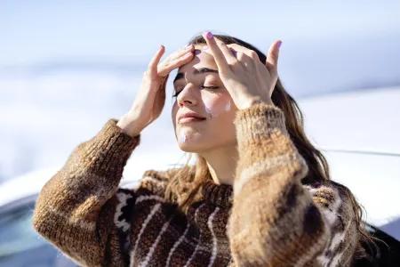 Eine junge Frau mit braunen Haaren trägt draußen bei sonnigem Winterwetter Gesichtscreme auf ihre Stirn auf. Sie trägt einen dicken Pullover und genießt sichtbar das Licht.
