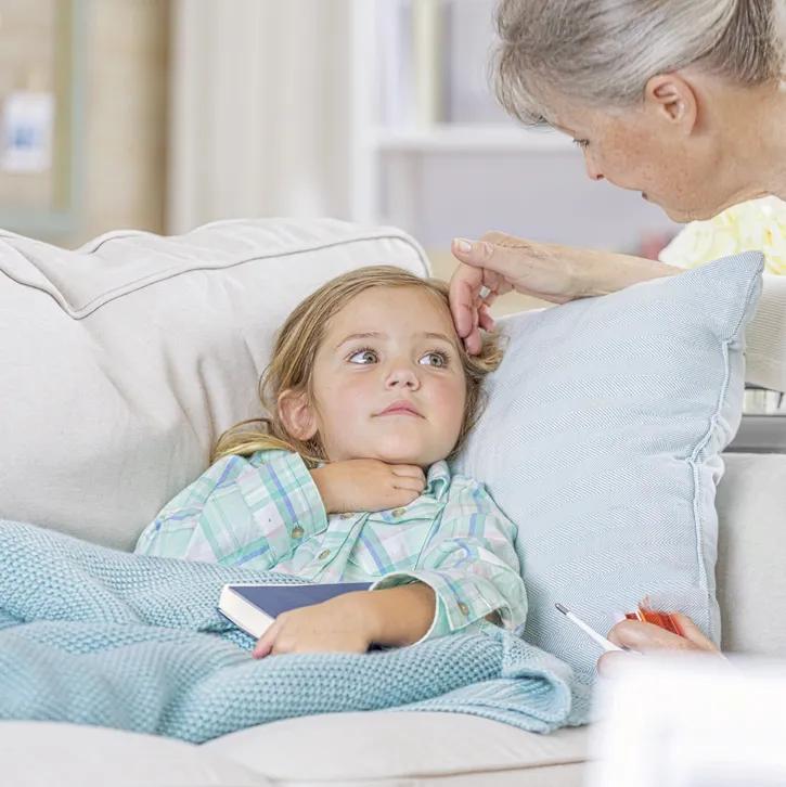 Ein kleines blondes M&auml;dchen liegt mit einem Buch auf dem Scho&szlig; krank auf dem Sofa, in Decke und Pyjama eingekuschelt. Eine &auml;ltere Frau f&uuml;hlt mit der Hand die Stirn des Kindes.