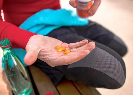 Frau in rotem Sportoberteil hält drei gelbliche Kapseln in der Hand, im Hintergrund Sportkleidung und eine Wasserflasche – Symbolbild für Nahrungsergänzungsmittel bei körperlicher Aktivität.