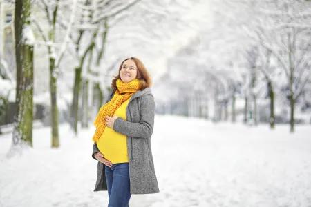 Schwangere Frau in Winterkleidung steht auf einem verschneiten Weg in einer Allee und l&auml;chelt zufrieden.