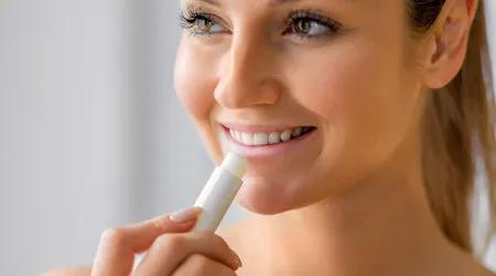 Eine junge Frau tr&auml;gt mit einem Pflegestift Lippenbalsam auf, w&auml;hrend sie l&auml;chelnd zur Seite schaut.