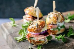 Drei kleine belegte Laugenbrötchen mit Lachs, Zwiebeln, Frischkäse und Rucola, angerichtet auf einem Holzbrett.
