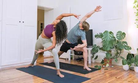 Zwei Personen in Sportkleidung führen gemeinsam eine Yoga- oder Pilates-Übung auf Matten in einem Wohnzimmer aus.