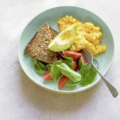 Teller mit Rührei, Vollkornbrot, Avocadoscheibe und kleinem Spinatsalat.