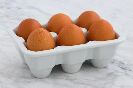 Sechs braune H&uuml;hnereier in einem wei&szlig;en Keramik-Eierhalter auf einem hellen Marmortisch &ndash; Symbol f&uuml;r proteinreiche Ern&auml;hrung und nat&uuml;rliche Lebensmittel.