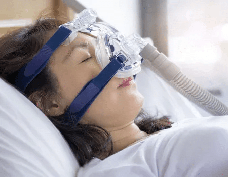 Frau liegt mit Schlafmaske und Atemtherapiegerät zur Behandlung von Schlafapnoe im Bett.