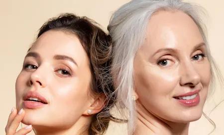 Zwei Frauen unterschiedlichen Alters mit makelloser Haut posieren nebeneinander und lächeln selbstbewusst in die Kamera.