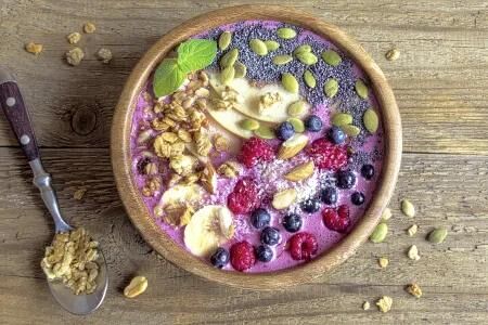 Bunte Smoothie-Bowl mit Bananen, Beeren, N&uuml;ssen und Samen in einer Holzschale auf rustikalem Holztisch