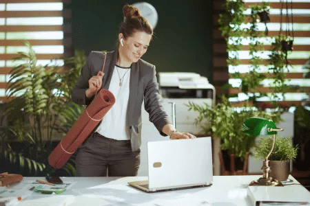 Businessfrau mit Yogamatte schließt Laptop im Büro und bereitet sich auf ihre Fitnessroutine in entspannter Arbeitsumgebung vor