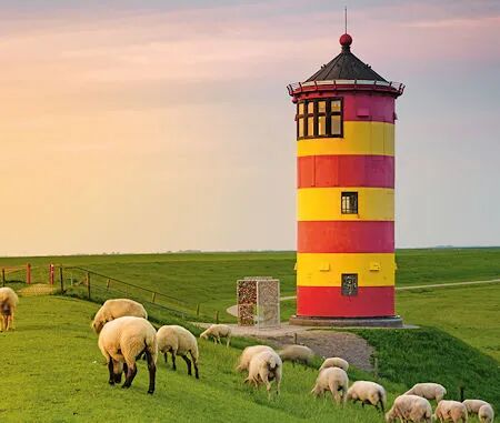 Ein rot-gelb gestreifter Leuchtturm auf einer grünen Wiese, umgeben von grasenden Schafen im warmen Licht des Sonnenuntergangs.