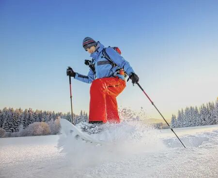 Person mit roter Hose beim Schneeschuhwandern in tiefem Schnee vor verschneiten Bäumen und blauen Himmel.