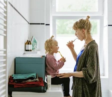 Eine Mutter und ihr kleines Kind stehen gemeinsam im Badezimmer und putzen sich nebeneinander die Zähne. Das Kind sitzt auf der Waschmaschine, beide schauen sich liebevoll an.
