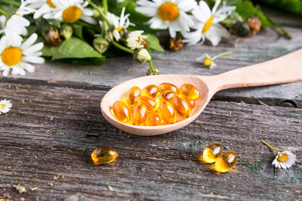 Ein Holzlöffel liegt auf einem rustikalen Holztisch, gefüllt mit gelben Vitamin-D-Kapseln. Drumherum liegen Kamillenblüten.