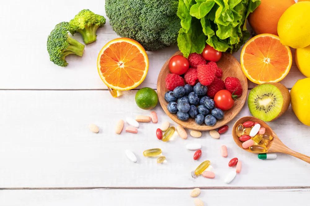 Obst, Gemüse, Vitaminpräparate und bunte Tabletten liegen auf einem weißen Tisch. Darunter Brokkoli, Orangen, Beeren, Kiwi und Nahrungsergänzungskapseln.