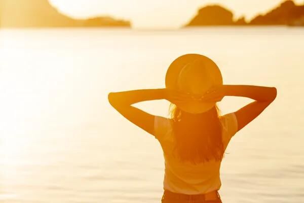 Eine Person steht bei Sonnenuntergang am Wasser, hält beide Arme an den Hut und blickt in die Sonne. Die Szene ist von warmem Licht durchflutet.